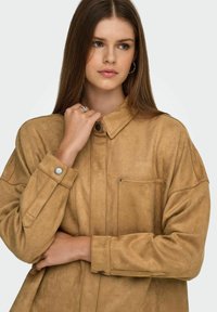 Tan kleurige suède shirtjas met een kraag met knopen, een borstzak en drukknoopmanchetten. Beschikt over een zachte textuur en een relaxte pasvorm.