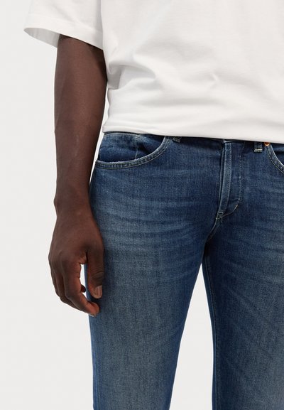 Jeans in denim blu scuro con un design aderente, caratterizzati da tasche frontali, leggera sfumatura e un lieve sbiadimento. Abbinati a una maglietta bianca semplice.