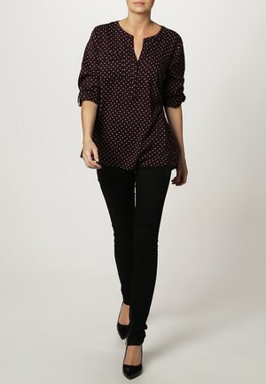 Mujer caminando con una blusa negra de manga larga con puntos blancos, pantalones negros y zapatos de tacón negro sobre un fondo liso.