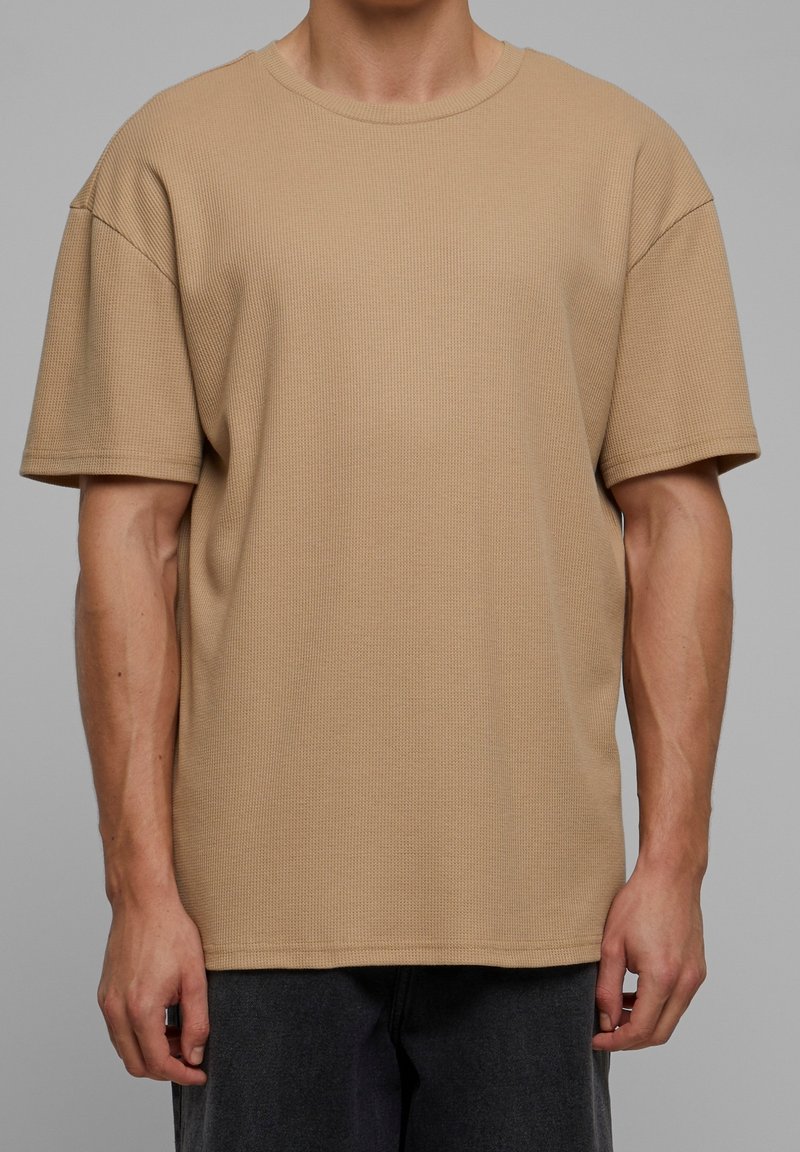 Homme portant un t-shirt beige uni à manches courtes en maille texturée et un pantalon foncé, debout devant un fond gris neutre.