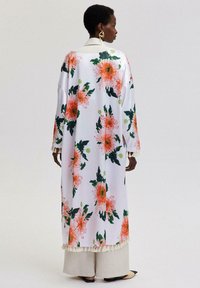 Blommig kimono i lätt tyg, med stora rosa blommor och gröna blad på en vit bakgrund, med fransdetaljer vid fållen.
