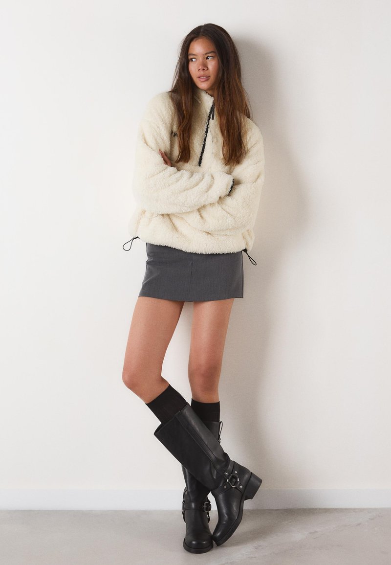 Mikina z flísu v krémovej farbe s vysokým golierom a nastaviteľnými šnúrkami, v kombinácii so sivou mini sukňou a čižmami z kože po kolená.