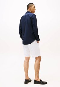 Camicia button-up blu navy con maniche lunghe, texture liscia e orlo curvo, abbinata a pantaloni corti bianchi e mocassini neri.