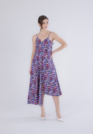 Kobieta stojąca z ręką na biodrze, ubrana w bezrękawnikową, wzorzystą sukienkę midi z czerwono-niebiesko-białym geometrycznym wzorem oraz białe szpilki.