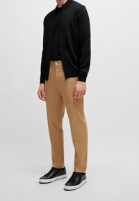 Černý pletený svetr na zip přes černé tričko, doplněný o světle béžové kalhoty a černé tenisky se šedými podrážkami. Jednoduchý design a hladká struktura.