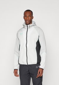 Veste légère à capuche avec un devant blanc, des côtés noirs et des accents menthe. Dispose d'une fermeture éclair sur le devant et de poignets élastiques pour un ajustement.