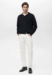 Black knitted pullover with a collar, white cotton pants, and black leather shoes. Model stands with hands in pockets against a plain background.