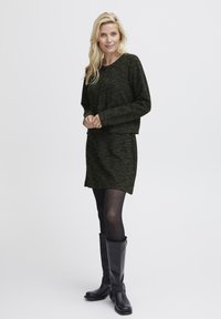 Czarna i zielona swetrowa sukienka o teksturowanej powierzchni z luźnym krojem, zestawiona z czarnymi botkami do kolan. Neutralne tło.