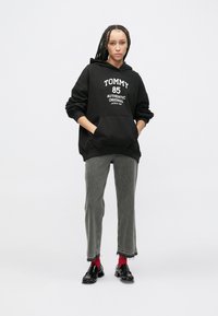 Čierna bavlnená mikina s kapucňou s potlačou "TOMMY 85", v kombinácii so sivými džínsami so strapcami na lemoch. Nosené s čiernymi topánkami a červenými ponožkami.