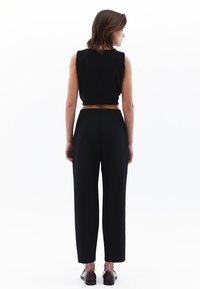 Zwarte mouwloze cropped top gecombineerd met hoge zwarte broeken. De broeken hebben een rechte snit en subtiele textuur; de schoenen zijn donkere leren loafers.