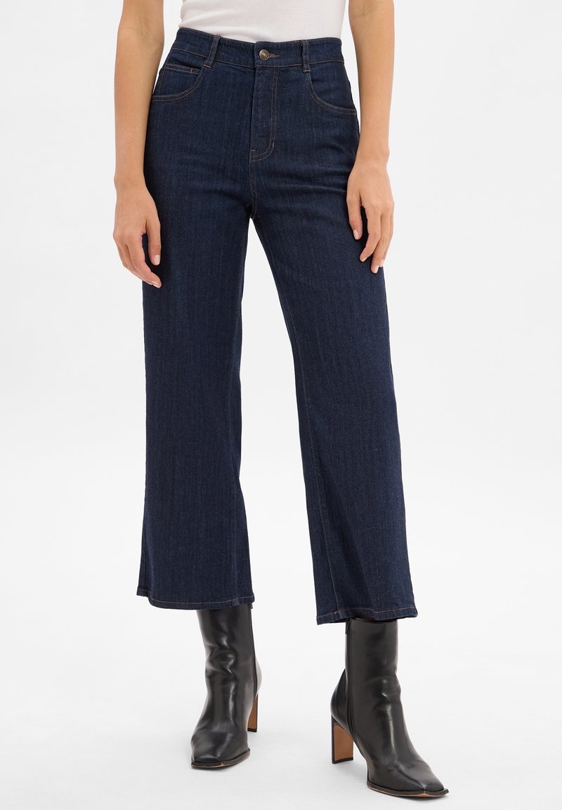 Ciemnoniebieskie dżinsy o szerokich nogawkach z wysokim stanem, wyposażone w przednie kieszenie i subtelne przeszycia. Połączone z czarnymi botkami na obcasie.