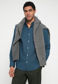 Mygtukais užsegama tamsiai žalsvo atspalvio marškiniai, ant jų per petis užmestas pilkas megztinis, derinamas su tamsiai žaliais kelnais. Lygus tekstūra.