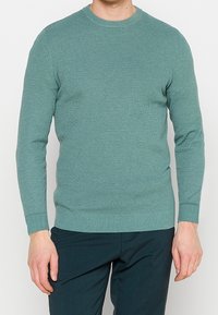 Jersey de punto turquesa con cuello redondo, mangas largas y puños acanalados. Tejido texturizado, estilo ajustado, combinado con pantalones verde oscuro.