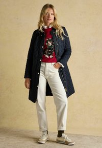 Námornícka prešívaná bunda so zlatými kovovými doplnkami, červený sveter s dizajnom psa, biele nohavice a svetlé tenisky s béžovými akcentmi.