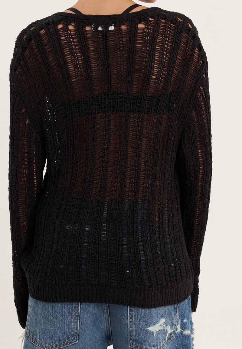 Vue de dos d'une personne portant un pull noir à maille large par-dessus un top noir et un jean bleu déchiré sur fond uni.