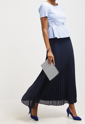 Femme portant un haut péplum bleu clair et une jupe longue plissée bleu marine, tenant une pochette rayée, marchant en escarpins bleus.