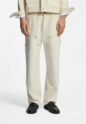 Dolna część ciała ubrana w kremowe spodnie o prostych nogawkach z przednimi kieszeniami naszywanymi oraz czarne wsuwane mokasyny, ręce luźno wzdłuż ciała.