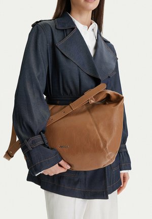 Mujer con chaqueta de mezclilla oscura y pantalones blancos, sosteniendo un gran bolso de cuero marrón con correa ajustable.