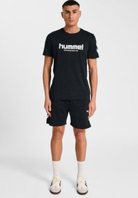 Schwarzes Baumwoll-T-Shirt mit "hummel"-Logo und grauen Sportshorts. Strukturierter Stoff, lässiger Schnitt, kombiniert mit weißen Socken und grauen Turnschuhen.