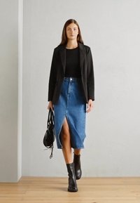 Schwarzer Blazer über einem schwarzen Oberteil, kombiniert mit einem hellblauen Jeansrock mit einem seitlichen Schlitz. Schwarze Stiefeletten und eine schwarze Tasche runden den Look ab.