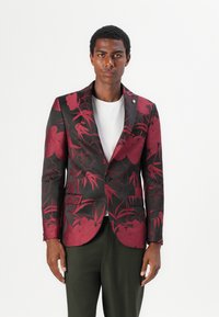 Schwarzer Blazer mit tiefrotem Blumenmuster. Ein-Knopf-Verschluss, Reverskragen, Taschen an der Vorderseite, über einem weißen T-Shirt getragen.
