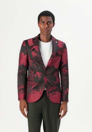 Schwarzer Blazer mit tiefrotem Blumenmuster. Ein-Knopf-Verschluss, Reverskragen, Taschen an der Vorderseite, über einem weißen T-Shirt getragen.