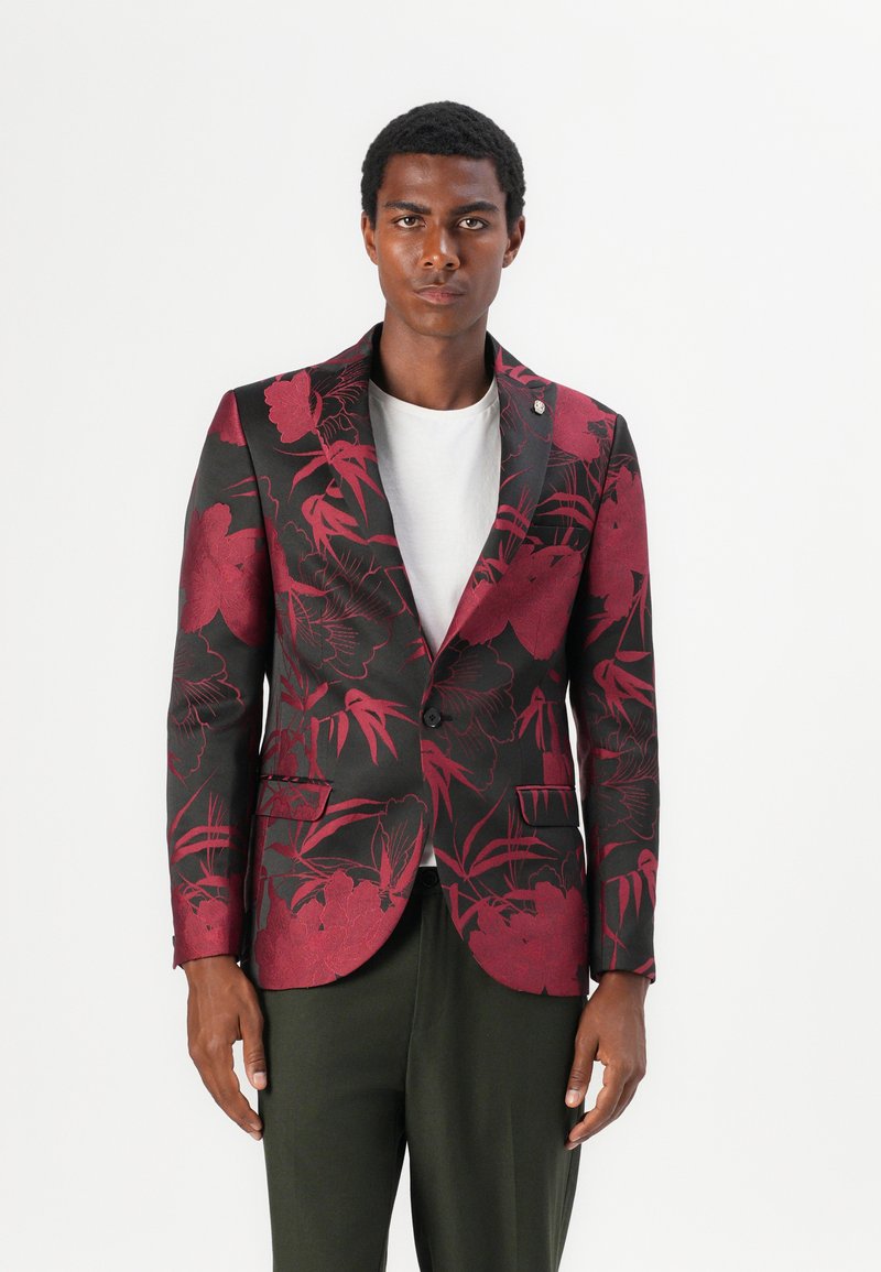 Schwarzer Blazer mit tiefrotem Blumenmuster. Ein-Knopf-Verschluss, Reverskragen, Taschen an der Vorderseite, über einem weißen T-Shirt getragen.