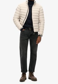 Giacca puffer color beige chiaro con motivo a cuciture orizzontali, maglione blu navy, jeans neri e stivali marroni con lacci. Dettagli notevoli sui polsini della giacca.
