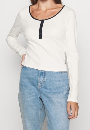 White long-sleeve top with a scoop neckline, featuring a black button placket and ribbed texture, paired with light blue denim jeans.