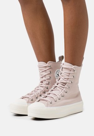 Persona che indossa sneakers alte con lacci di colore rosa chiaro, suole bianche spesse e punte tonde, in piedi contro uno sfondo semplice.