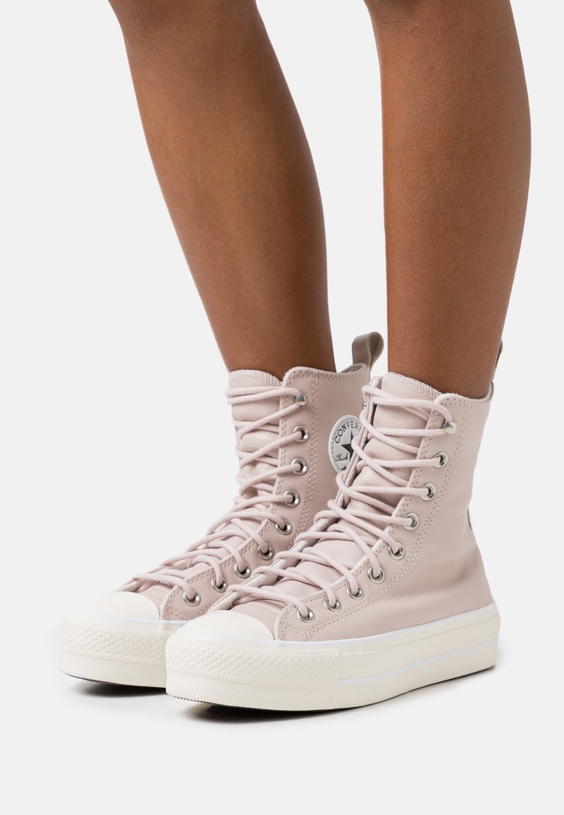 Personne portant des baskets montantes à lacets de couleur rose clair avec des semelles épaisses blanches et des embouts ronds, debout devant un fond uni.