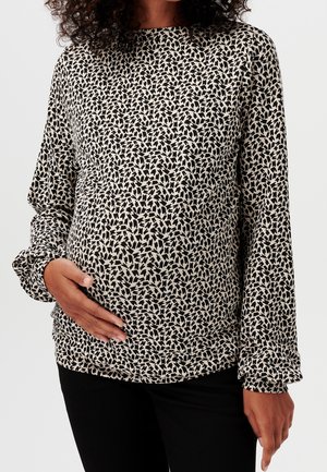 Nainen, jolla on mustavalkokuvioitu pitkähihainen paita, pitää toista kättään raskausvatsallaan, mustat housut näkyvissä.