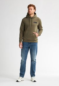Felpa verde oliva con cordini regolabili, tasca frontale e piccolo logo nero. Abbinata a jeans dritti blu chiaro e sneakers bianche.