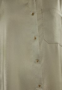 Satin-olivgrünes Hemd mit Knopfleiste, sechs braunen Knöpfen und einer linken Brusttasche, das einen leichten Glanz und eine glatte Textur aufweist.