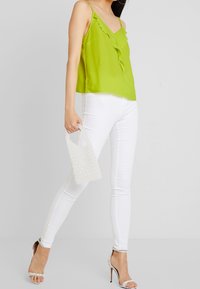 Top sin mangas verde lima con detalle de volantes, jeans blancos ajustados y un bolso transparente con textura; combinados con tacones de tiras con detalles de brillantes.