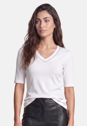Frau mit langen dunklen Haaren, die ein weißes kurzärmeliges V-Ausschnitt-Oberteil und schwarze hoch taillierte Lederhose trägt und leicht zur Seite schaut.