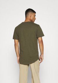 Olive green short-sleeve t-shirt made of soft cotton with a curved hem, paired with light beige trousers. Back view of the fit.