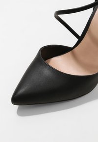 Chaussure noire à bout pointu en cuir texturé avec un intérieur nude et de fines lanières croisées près de la cheville.