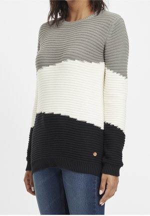 Kobieta ubrana w prążkowany sweter z dzianiny z poziomymi blokami kolorów w odcieniach szarości, bieli i czerni, w połączeniu z niebieskimi dżinsami.