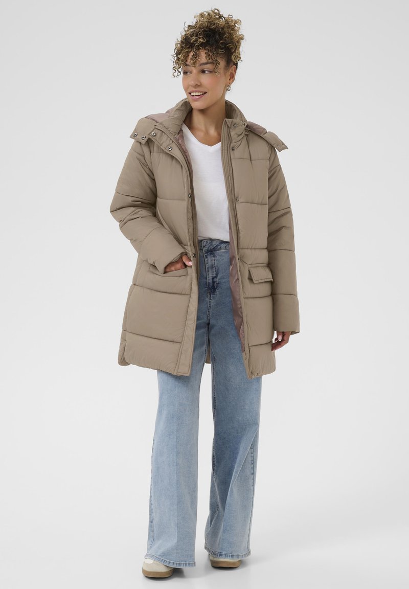 Doudoune beige avec une capuche, présentant une texture matelassée et des poches latérales, portée sur un t-shirt blanc et un jean flare bleu clair.