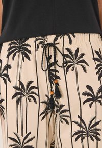 Lichtgekleurde broek met een zwart palmbomenpatroon, elastische tailleband met een zwarte trekkoord en decoratieve kwastjes, en houten kralen.