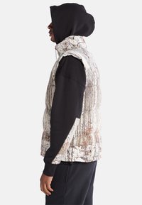 Gestepptes Weste mit Baumrindenmuster und hohem Kragen, darunter ein schwarzes Langarmshirt und eine Kapuze. Schwarze Jogginghosen sind sichtbar.