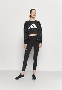 Felpa crop nera con motivo screziato bianco e grande logo Adidas, abbinata a leggings neri con strisce laterali bianche e sneakers nere.