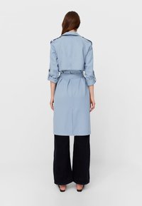 Jasnoniebieski trencz z paskiem w talii, rękawami długości trzy czwarte i klasycznym kołnierzem; zestawiony z czarnymi szerokimi spodniami.