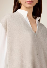Beige gestrickte Weste mit V-Ausschnitt, die einen dezenten Glanz aufweist, kurze Ärmel hat und vorne über einem weißen Hemd mit einem einzigen hellblauen Knopf versehen ist.