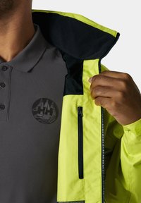 Chaqueta impermeable amarilla brillante con acentos negros, bolsillo con cremallera, llevada sobre una camiseta polo texturizada gris con un emblema negro.