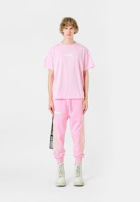 Camiseta de algodón rosa de gran tamaño con una etiqueta rectangular en la parte frontal, combinada con pantalones de chándal rosas que presentan diseños de plumas en relieve y cordón.