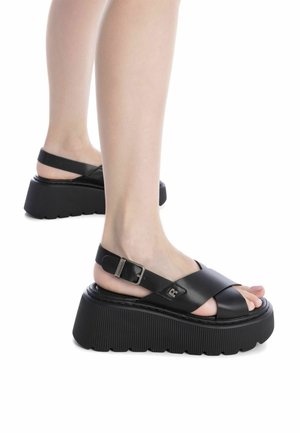 Sandalias de plataforma negras con tiras cruzadas anchas, correa ajustable en el tobillo y suela texturizada con estrías. Fabricadas en cuero suave con detalles de metal.