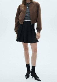 Veste bomber courte marron, chemise grise, jupe plissée noire, chaussettes noires, escarpins noirs à talons pointus. Texture : veste lisse, jupe en tissu.