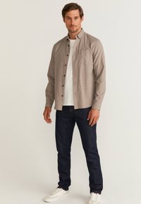 Lichtbruine overhemd met knopen en een zak, gedragen over een wit t-shirt. Gecombineerd met donkerblauwe jeans en witte sneakers. Casual ontwerp.